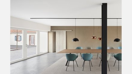 Heller, reduzierter Besprechungsraum mit langem Holztisch und bunten Stühlen: große Fenster nach außen links, Schiebetür aus Holz an der Rückwand, mehrere hängende Pendelleuchten (schwarz und kupferfarben) und eine dunkle Stütze mittig im Bild.