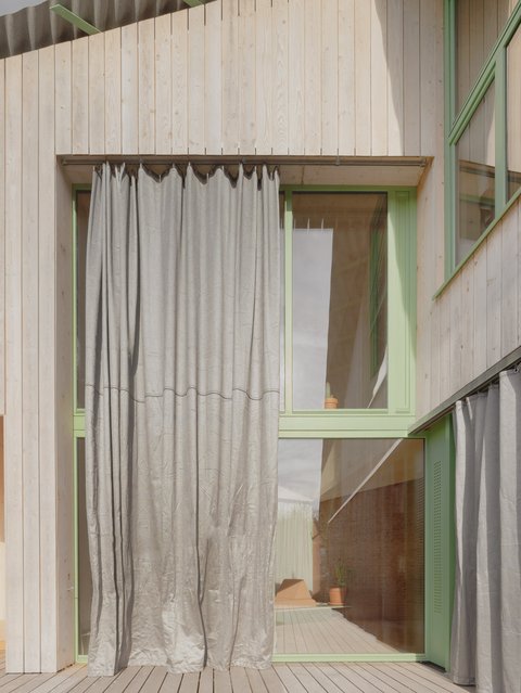  "Innenbereich mit heller Holzfassade und grünem Fensterrahmen. Weiße Vorhänge verdecken teilweise die Glasfläche. Holzterrasse davor."