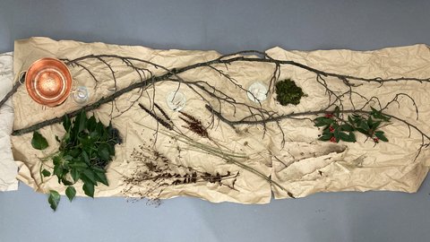 Auf braunem Packpapier sind akribisch Naturmaterialien angeordnet: ein kleiner Kupferkessel, Zweige, Efeu, Moos, Rindenstücke und kleine Gefäße mit getrockneten Pflanzenteilen.