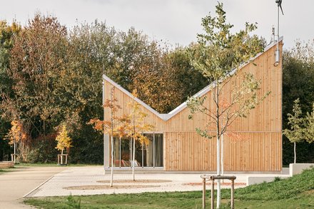 Kleiner Holzpavillon mit geknicktem Dach in einer Parklandschaft; Blumenbeet vorn, herbstliche Bäume hinten, große Schiebetüren.