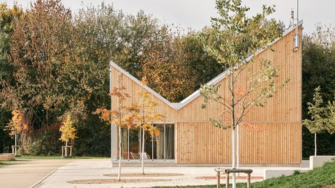 Kleiner Holzpavillon mit geknicktem Dach in einer Parklandschaft; Blumenbeet vorn, herbstliche Bäume hinten, große Schiebetüren.