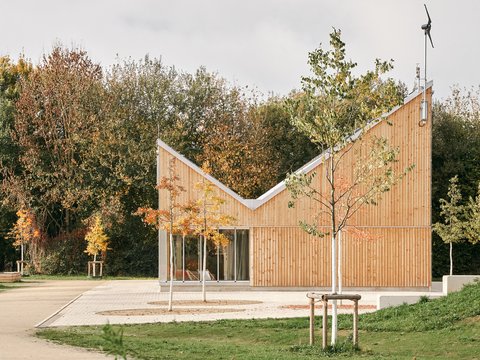 Kleiner Holzpavillon mit geknicktem Dach in einer Parklandschaft; Blumenbeet vorn, herbstliche Bäume hinten, große Schiebetüren.