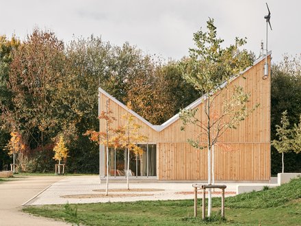 Kleiner Holzpavillon mit geknicktem Dach in einer Parklandschaft; Blumenbeet vorn, herbstliche Bäume hinten, große Schiebetüren.