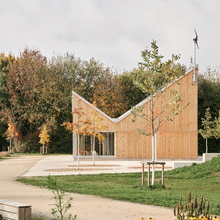Kleiner Holzpavillon mit geknicktem Dach in einer Parklandschaft; Blumenbeet vorn, herbstliche Bäume hinten, große Schiebetüren.
