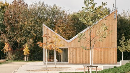 Kleiner Holzpavillon mit geknicktem Dach in einer Parklandschaft; Blumenbeet vorn, herbstliche Bäume hinten, große Schiebetüren.