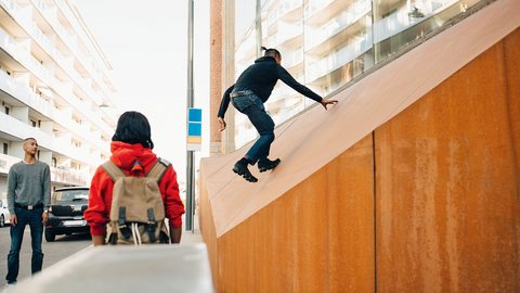 "Person in dunklem Oberteil und Jeans springt auf einer beige-orangefarbenen geneigten Holzfläche. Im Hintergrund stehen zwei weitere Personen in einem Innenhof mit modernen Wohnblöcken und großen Glasfenstern."