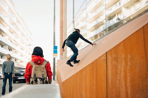 "Person in dunklem Oberteil und Jeans springt auf einer beige-orangefarbenen geneigten Holzfläche. Im Hintergrund stehen zwei weitere Personen in einem Innenhof mit modernen Wohnblöcken und großen Glasfenstern."