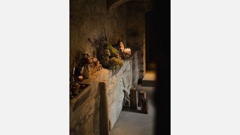 Foto eines rustikalen Yoga- oder Meditationsraums. Sonnenlicht fällt durch ein verborgenes Fenster auf der linken Seite. Decken und Kissen liegen gefaltet auf dem Boden, und ein Gong mit chinesischen Schriftzeichen hängt an einem Holzbalken in der Mitte. Makramee-Seile hängen auf beiden Seiten.  image_27.png