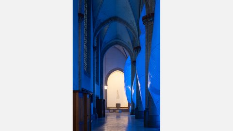 Blick durch das Kirchenschiff mit hohen Spitzbögen und blau beleuchteten Stoffinstallationen. Im Hintergrund öffnet sich ein heller Altarraum.