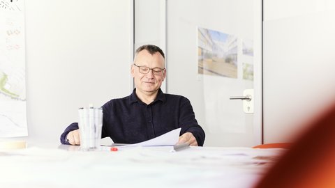 Ein älterer Mann sitzt an einem Tisch in einem Büro und liest ein Dokument. Vor ihm stehen Stifte in einem Becher. Der Raum ist hell, mit schlichten Wänden und einer Tür im Hintergrund.