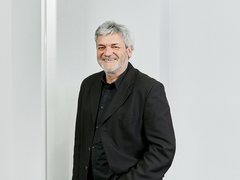 "Porträtfoto eines älteren Mannes mit grauem Haar und Bart, der freundlich lächelt. Er trägt einen schwarzen Blazer über einem schwarzen Hemd. Er steht vor einem hellen, minimalistischen Hintergrund mit weißer Wand. Professionelle Headshot-Fotografie."