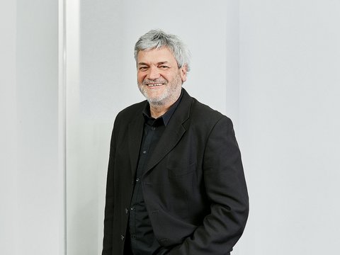 "Porträtfoto eines älteren Mannes mit grauem Haar und Bart, der freundlich lächelt. Er trägt einen schwarzen Blazer über einem schwarzen Hemd. Er steht vor einem hellen, minimalistischen Hintergrund mit weißer Wand. Professionelle Headshot-Fotografie."