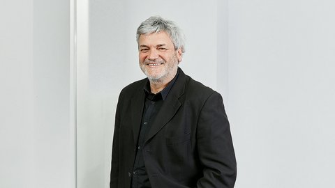 "Porträtfoto eines älteren Mannes mit grauem Haar und Bart, der freundlich lächelt. Er trägt einen schwarzen Blazer über einem schwarzen Hemd. Er steht vor einem hellen, minimalistischen Hintergrund mit weißer Wand. Professionelle Headshot-Fotografie."