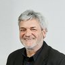 "Porträtfoto eines älteren Mannes mit grauem Haar und Bart, der freundlich lächelt. Er trägt einen schwarzen Blazer über einem schwarzen Hemd. Er steht vor einem hellen, minimalistischen Hintergrund mit weißer Wand. Professionelle Headshot-Fotografie."