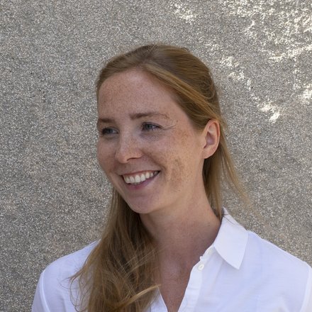 Porträt einer Person mit langen rötlichen Haaren im weißen Hemd, lächelnd und nach links blickend; Hintergrund: helle, grob verputzte Wand mit schrägem Licht.