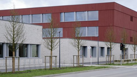 Außenansicht eines modernen, mehrstöckigen Gebäudes mit auffälliger roter Holzfassade im Obergeschoss und hellem Klinker im Erdgeschoss.