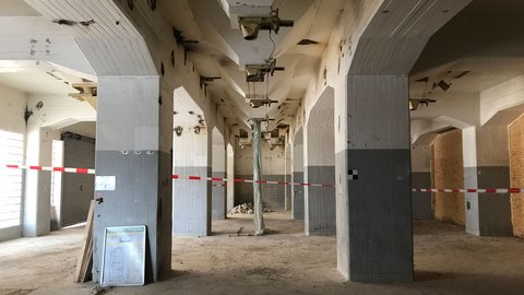  Leerer Industrie-Innenraum im Umbau: Reihen grauer Betonstützen, rot‑weißes Absperrband sperrt den Mittelgang, hinten Bauschutt; vorn lehnt ein Bauplan an einer Säule.