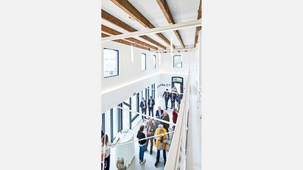Vogelperspektive auf einen hellen, zweigeschossigen Veranstaltungsraum mit sichtbaren Holzbalken, großen Fenstern und mehreren stehenden Gästen bei einem Empfang.