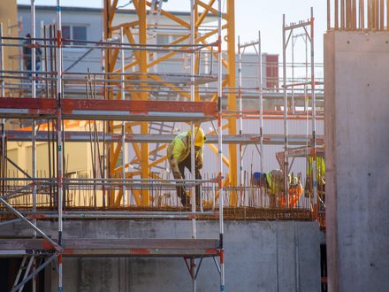 Bauarbeiter in gelber Schutzkleidung arbeiten auf Gerüsten an einer Betonplatte mit herausragender Bewehrung; im Hintergrund ein gelber Turmkran und Baustellenstrukturen im warmen Gegenlicht.