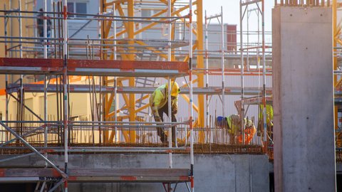 Bauarbeiter in gelber Schutzkleidung arbeiten auf Gerüsten an einer Betonplatte mit herausragender Bewehrung; im Hintergrund ein gelber Turmkran und Baustellenstrukturen im warmen Gegenlicht.