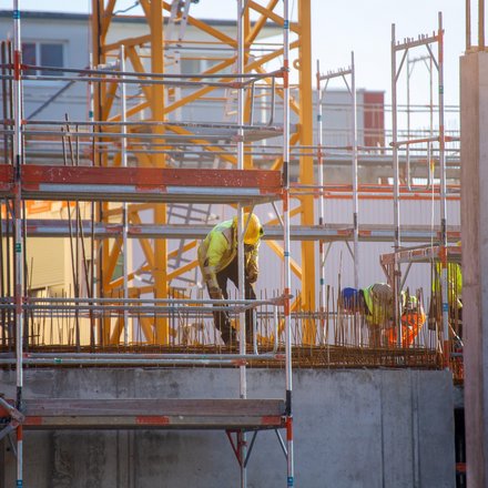 Bauarbeiter in gelber Schutzkleidung arbeiten auf Gerüsten an einer Betonplatte mit herausragender Bewehrung; im Hintergrund ein gelber Turmkran und Baustellenstrukturen im warmen Gegenlicht.
