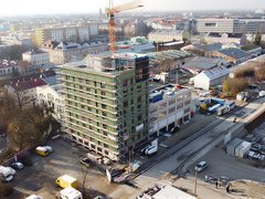 Das Bild zeigt eine Luftaufnahme einer städtischen Baustelle. Im Zentrum steht ein mehrgeschossiges Gebäude, das von einem Gerüst umgeben ist und sich im Bau befindet. Ein Baukran ragt über das Gebäude hinaus. Rundherum sind Straßen, weitere Gebäude, Lagerflächen und Baumaterialien zu sehen. Die städtische Umgebung ist dicht bebaut, was den Bau im urbanen Kontext deutlich macht. Zweck des Bildes ist es, ein größeres Bauprojekt im Stadtraum zu zeigen und den Maßstab sowie die Komplexität zirkulären Bauens im urbanen Umfeld zu verdeutlichen.