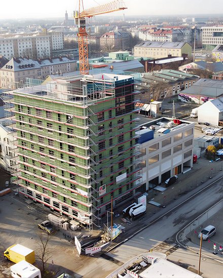 Das Bild zeigt eine Luftaufnahme einer städtischen Baustelle. Im Zentrum steht ein mehrgeschossiges Gebäude, das von einem Gerüst umgeben ist und sich im Bau befindet. Ein Baukran ragt über das Gebäude hinaus. Rundherum sind Straßen, weitere Gebäude, Lagerflächen und Baumaterialien zu sehen. Die städtische Umgebung ist dicht bebaut, was den Bau im urbanen Kontext deutlich macht. Zweck des Bildes ist es, ein größeres Bauprojekt im Stadtraum zu zeigen und den Maßstab sowie die Komplexität zirkulären Bauens im urbanen Umfeld zu verdeutlichen.