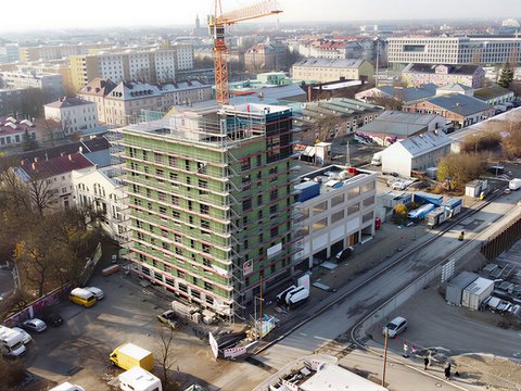 Das Bild zeigt eine Luftaufnahme einer städtischen Baustelle. Im Zentrum steht ein mehrgeschossiges Gebäude, das von einem Gerüst umgeben ist und sich im Bau befindet. Ein Baukran ragt über das Gebäude hinaus. Rundherum sind Straßen, weitere Gebäude, Lagerflächen und Baumaterialien zu sehen. Die städtische Umgebung ist dicht bebaut, was den Bau im urbanen Kontext deutlich macht. Zweck des Bildes ist es, ein größeres Bauprojekt im Stadtraum zu zeigen und den Maßstab sowie die Komplexität zirkulären Bauens im urbanen Umfeld zu verdeutlichen.
