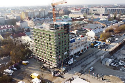 Das Bild zeigt eine Luftaufnahme einer städtischen Baustelle. Im Zentrum steht ein mehrgeschossiges Gebäude, das von einem Gerüst umgeben ist und sich im Bau befindet. Ein Baukran ragt über das Gebäude hinaus. Rundherum sind Straßen, weitere Gebäude, Lagerflächen und Baumaterialien zu sehen. Die städtische Umgebung ist dicht bebaut, was den Bau im urbanen Kontext deutlich macht. Zweck des Bildes ist es, ein größeres Bauprojekt im Stadtraum zu zeigen und den Maßstab sowie die Komplexität zirkulären Bauens im urbanen Umfeld zu verdeutlichen.