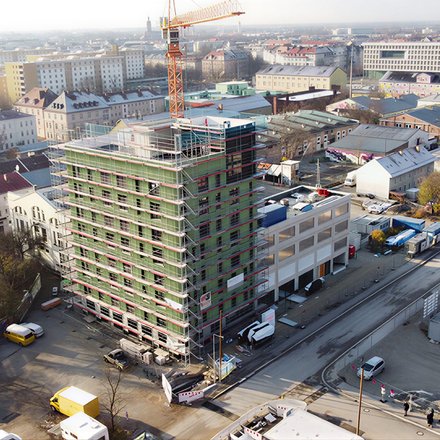 Das Bild zeigt eine Luftaufnahme einer städtischen Baustelle. Im Zentrum steht ein mehrgeschossiges Gebäude, das von einem Gerüst umgeben ist und sich im Bau befindet. Ein Baukran ragt über das Gebäude hinaus. Rundherum sind Straßen, weitere Gebäude, Lagerflächen und Baumaterialien zu sehen. Die städtische Umgebung ist dicht bebaut, was den Bau im urbanen Kontext deutlich macht. Zweck des Bildes ist es, ein größeres Bauprojekt im Stadtraum zu zeigen und den Maßstab sowie die Komplexität zirkulären Bauens im urbanen Umfeld zu verdeutlichen.