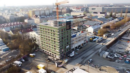 Das Bild zeigt eine Luftaufnahme einer städtischen Baustelle. Im Zentrum steht ein mehrgeschossiges Gebäude, das von einem Gerüst umgeben ist und sich im Bau befindet. Ein Baukran ragt über das Gebäude hinaus. Rundherum sind Straßen, weitere Gebäude, Lagerflächen und Baumaterialien zu sehen. Die städtische Umgebung ist dicht bebaut, was den Bau im urbanen Kontext deutlich macht. Zweck des Bildes ist es, ein größeres Bauprojekt im Stadtraum zu zeigen und den Maßstab sowie die Komplexität zirkulären Bauens im urbanen Umfeld zu verdeutlichen.