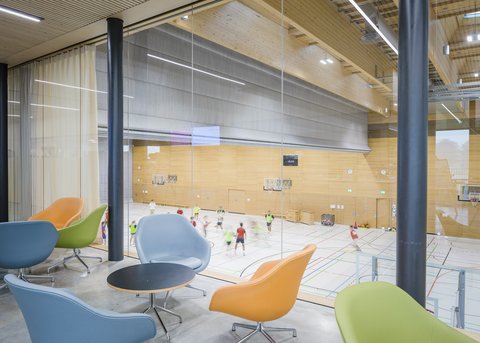 Loungebereich mit Blick in eine Sporthalle durch raumhohe Verglasung.