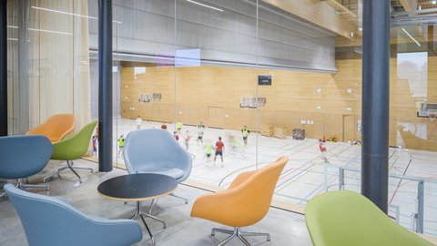 Loungebereich mit Blick in eine Sporthalle durch raumhohe Verglasung.