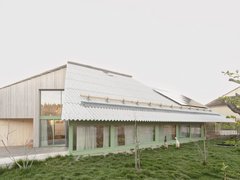 "Modernes Wohngebäude mit geneigtem Wellblechdach und grünem Fensterrahmen. Das Gebäude hat eine helle Holzfassade, große Glasflächen und ist umgeben von Rasenfläche mit jungen Bäumen. Im Hintergrund Wald."