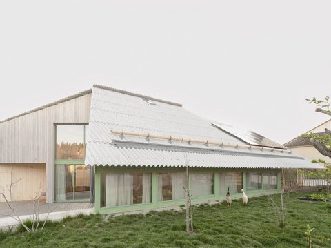 "Modernes Wohngebäude mit geneigtem Wellblechdach und grünem Fensterrahmen. Das Gebäude hat eine helle Holzfassade, große Glasflächen und ist umgeben von Rasenfläche mit jungen Bäumen. Im Hintergrund Wald."