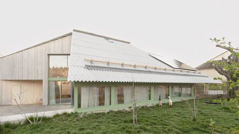 "Modernes Wohngebäude mit geneigtem Wellblechdach und grünem Fensterrahmen. Das Gebäude hat eine helle Holzfassade, große Glasflächen und ist umgeben von Rasenfläche mit jungen Bäumen. Im Hintergrund Wald."