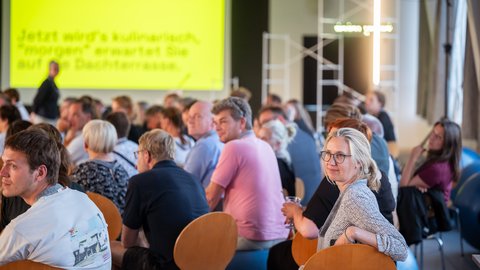 „Ein gut besuchtes Veranstaltungspublikum: Einige Teilnehmer blicken lächelnd zurück, während auf einer großen gelben Leinwand im Hintergrund der kulinarische Teil auf der Dachterrasse angekündigt wird.“