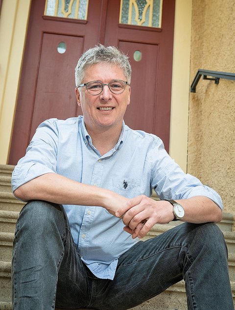 Porträt einer lächelnden Person mittleren Alters mit kurzen grauen Haaren und Brille, die in einem hellblauen Hemd mit hochgekrempelten Ärmeln und Jeans entspannt auf Außentreppen vor einer rotbraunen Haustür sitzt; die Hände sind locker übereinandergelegt und eine Armbanduhr ist sichtbar.