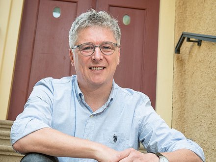 Porträt einer lächelnden Person mittleren Alters mit kurzen grauen Haaren und Brille, die in einem hellblauen Hemd mit hochgekrempelten Ärmeln und Jeans entspannt auf Außentreppen vor einer rotbraunen Haustür sitzt; die Hände sind locker übereinandergelegt und eine Armbanduhr ist sichtbar.