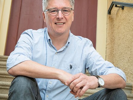 Porträt einer lächelnden Person mittleren Alters mit kurzen grauen Haaren und Brille, die in einem hellblauen Hemd mit hochgekrempelten Ärmeln und Jeans entspannt auf Außentreppen vor einer rotbraunen Haustür sitzt; die Hände sind locker übereinandergelegt und eine Armbanduhr ist sichtbar.