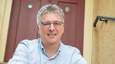 Porträt einer lächelnden Person mittleren Alters mit kurzen grauen Haaren und Brille, die in einem hellblauen Hemd mit hochgekrempelten Ärmeln und Jeans entspannt auf Außentreppen vor einer rotbraunen Haustür sitzt; die Hände sind locker übereinandergelegt und eine Armbanduhr ist sichtbar.
