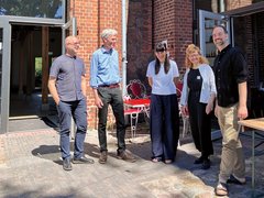 „Drei Männer und zwei Frauen stehen lächelnd in einem sonnigen Innenhof zwischen sanierten Backsteingebäuden. Über ihnen hängen Lichterketten und eine kleine Spiegelkugel.“