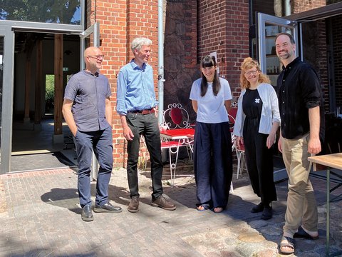 „Drei Männer und zwei Frauen stehen lächelnd in einem sonnigen Innenhof zwischen sanierten Backsteingebäuden. Über ihnen hängen Lichterketten und eine kleine Spiegelkugel.“