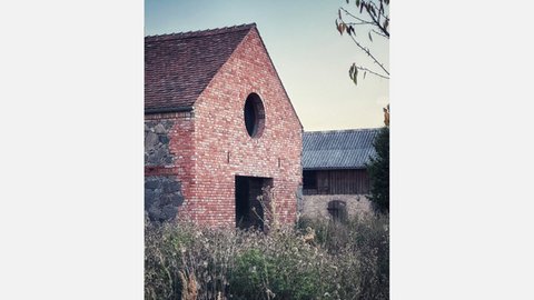 Giebel einer Ziegel‑ und Feldsteinscheune mit rundem Fenster und großer Toröffnung; wild bewachsener Vordergrund, Nachbargebäude im Hintergrund.