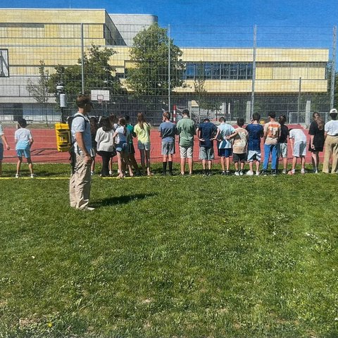Eine Schulklasse steht in einer Reihe auf einer Rasenfläche vor einem Sportplatz und einem Schulgebäude, beobachtet von einem Mann mit gelbem Rucksack.