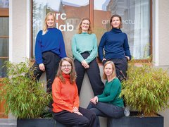"Fünf Frauen posieren vor einem modernen Bürogebäude mit der Aufschrift 'arc lab'. Hinten stehen drei Frauen: eine mit blondem Haar in blauem Pullover, eine mit rötlichem Haar und Brille in mintgrünem Pullover, eine mit braunem Haar in dunklem Rollkragenpullover. Vorne sitzen zwei Frauen: eine mit rötlichem Haar und Brille in rotem Rollkragenpullover, eine mit blondem Haar in grünem Pullover. Links und rechts sind Grünpflanzen sichtbar."