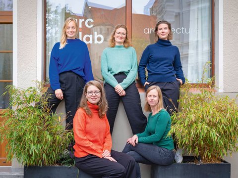 "Fünf Frauen posieren vor einem modernen Bürogebäude mit der Aufschrift 'arc lab'. Hinten stehen drei Frauen: eine mit blondem Haar in blauem Pullover, eine mit rötlichem Haar und Brille in mintgrünem Pullover, eine mit braunem Haar in dunklem Rollkragenpullover. Vorne sitzen zwei Frauen: eine mit rötlichem Haar und Brille in rotem Rollkragenpullover, eine mit blondem Haar in grünem Pullover. Links und rechts sind Grünpflanzen sichtbar."