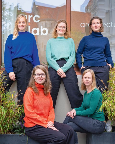 "Fünf Frauen posieren vor einem modernen Bürogebäude mit der Aufschrift 'arc lab'. Hinten stehen drei Frauen: eine mit blondem Haar in blauem Pullover, eine mit rötlichem Haar und Brille in mintgrünem Pullover, eine mit braunem Haar in dunklem Rollkragenpullover. Vorne sitzen zwei Frauen: eine mit rötlichem Haar und Brille in rotem Rollkragenpullover, eine mit blondem Haar in grünem Pullover. Links und rechts sind Grünpflanzen sichtbar."