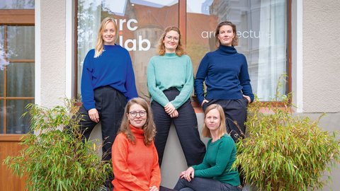 "Fünf Frauen posieren vor einem modernen Bürogebäude mit der Aufschrift 'arc lab'. Hinten stehen drei Frauen: eine mit blondem Haar in blauem Pullover, eine mit rötlichem Haar und Brille in mintgrünem Pullover, eine mit braunem Haar in dunklem Rollkragenpullover. Vorne sitzen zwei Frauen: eine mit rötlichem Haar und Brille in rotem Rollkragenpullover, eine mit blondem Haar in grünem Pullover. Links und rechts sind Grünpflanzen sichtbar."