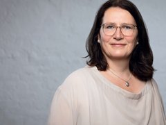 "Porträt einer Frau mittleren Alters mit schulterlangenm schwarzem Haar. Sie trägt eine Brille mit transparentem Rahmen und hat einen ruhigen, freundlichen Gesichtsausdruck. Um ihren Hals trägt sie eine silberne Kette mit einem ovalen Anhänger. In ihren Ohren sind diskrete Perlenohrringe sichtbar. Sie trägt ein beiges, cremefarbenes T-Shirt mit rundem Ausschnitt. Der Hintergrund ist neutral grau gehalten, was den Fokus auf die Person lenkt. Die Beleuchtung ist weich und gleichmäßig."